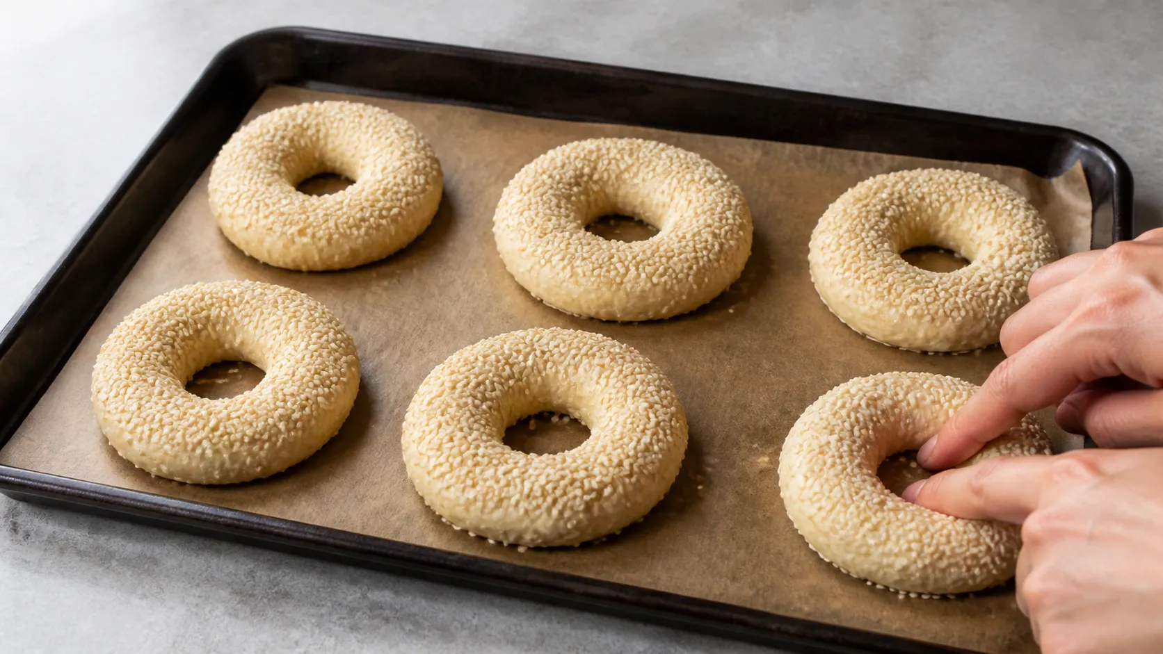 Hände formen sechs Keto-Bagel-Teigringe mit Sesam auf einem mit Backpapier belegten Backblech