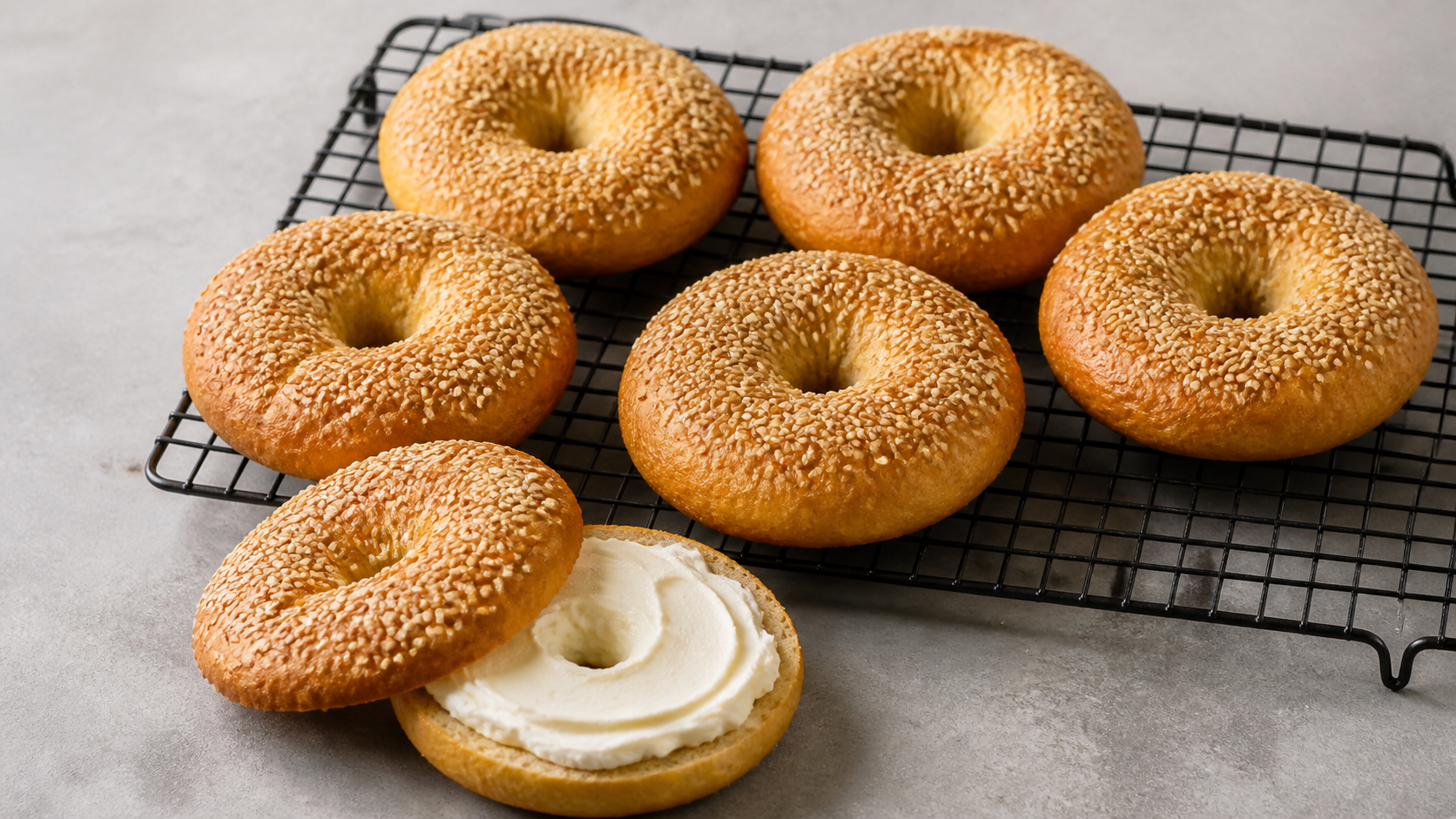 Sechs goldbraune Bagels mit Sesamkruste auf einem Kuchengitter, einer davon aufgeschnitten und mit Frischkäse bestrichen