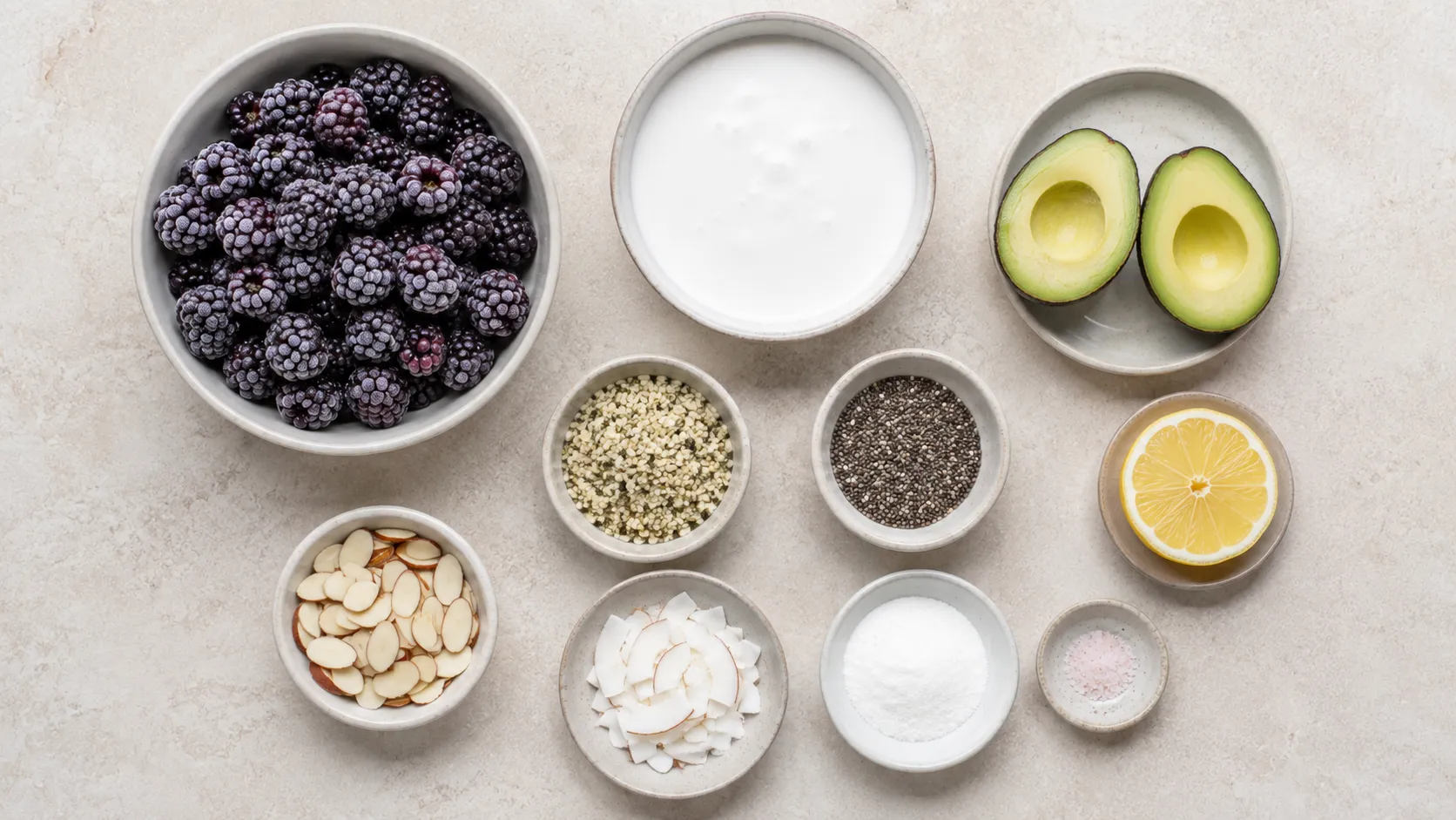 Zutaten für eine Keto-Smoothie-Bowl: gefrorene Brombeeren, kleine halbierte Avocado, Schale Kokosmilch, Hanfsamen, Chiasamen, Mandelblättchen, Kokoschips und Zitrone