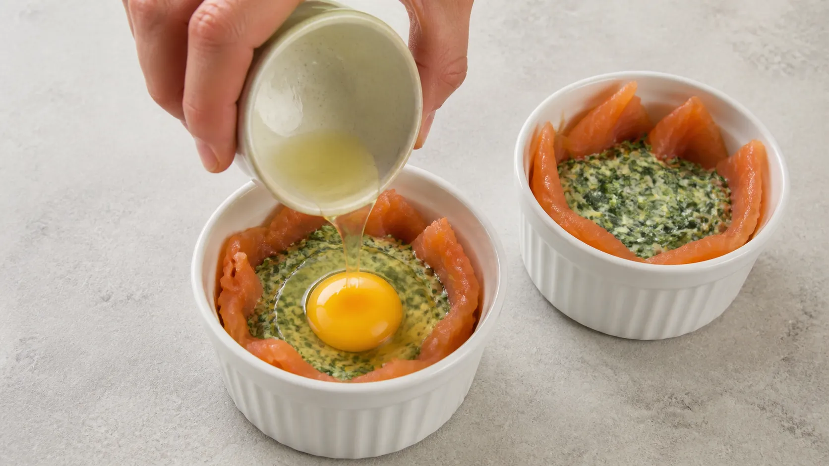 Auflaufförmchen mit Rahmspinat und Lachs, in die gerade ein rohes Ei gleiten gelassen wird