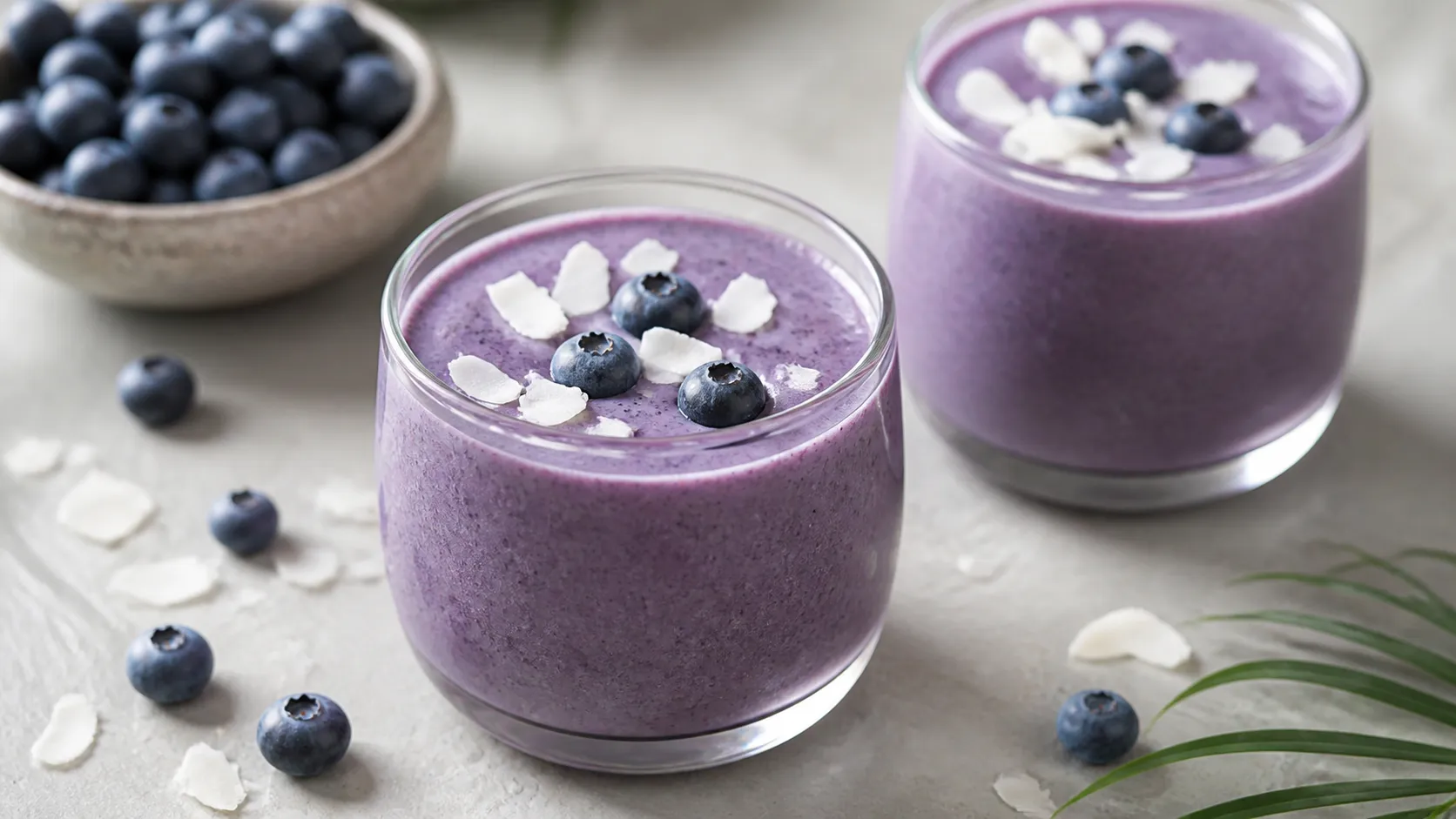 Cremiger violetter Keto-Heidelbeer-Kokos-Lassi in zwei Gläsern mit Kokosflocken und frischen Heidelbeeren