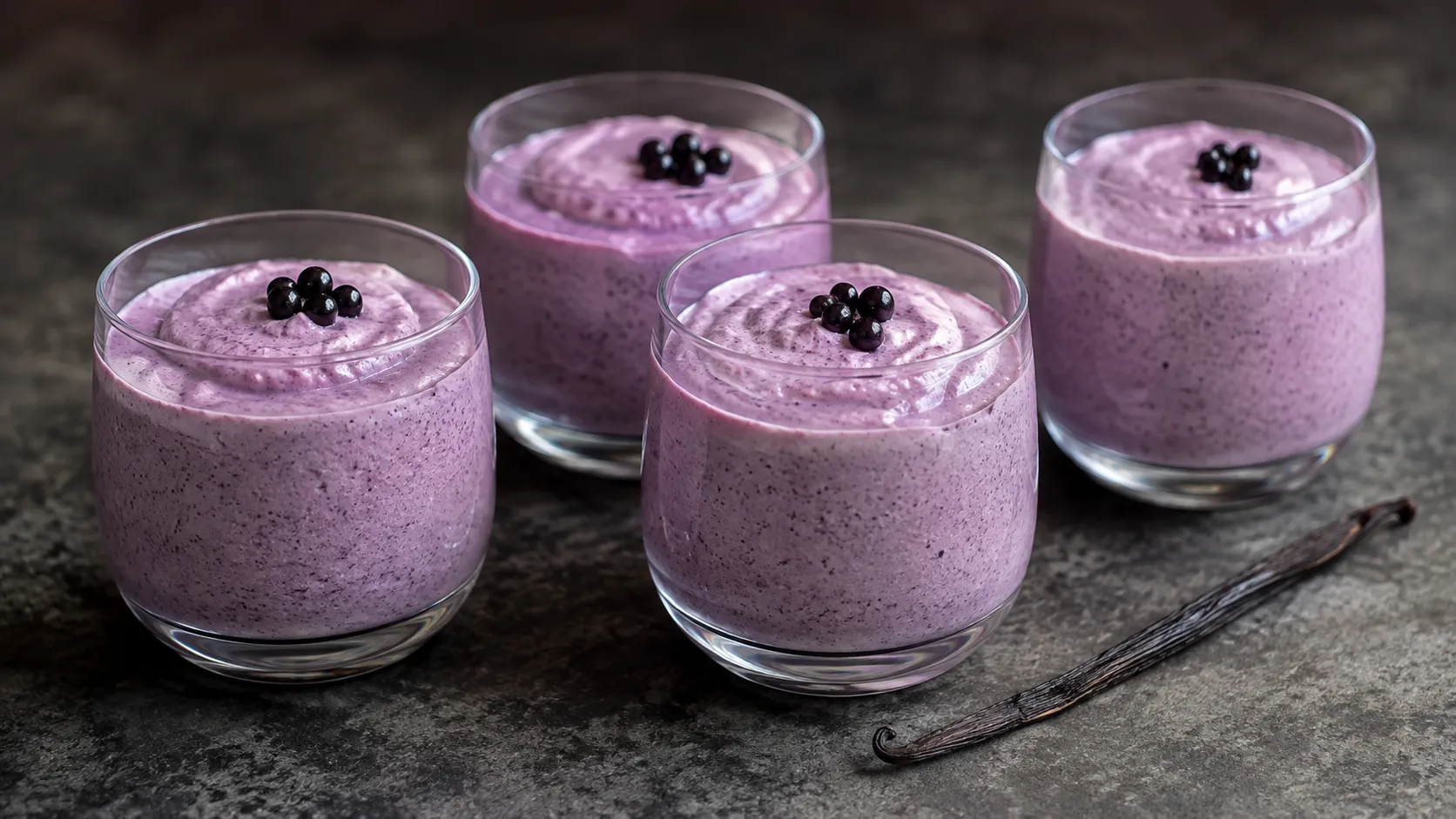 Vier Gläser mit hell-violetter cremiger Holunderbeer-Mousse, dekoriert mit dunklen Beeren und Vanilleschote