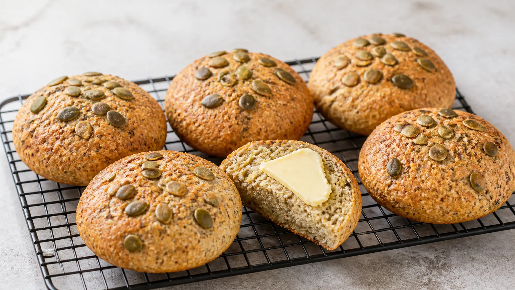 Sechs goldbraune Keto-Hüttenkäse-Brötchen mit Kürbiskernen auf einem Gitterrost, eines aufgeschnitten und mit Butter bestrichen