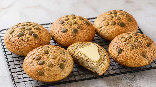 Sechs goldbraune Keto-Hüttenkäse-Brötchen mit Kürbiskernen auf einem Gitterrost, eines aufgeschnitten und mit Butter bestrichen