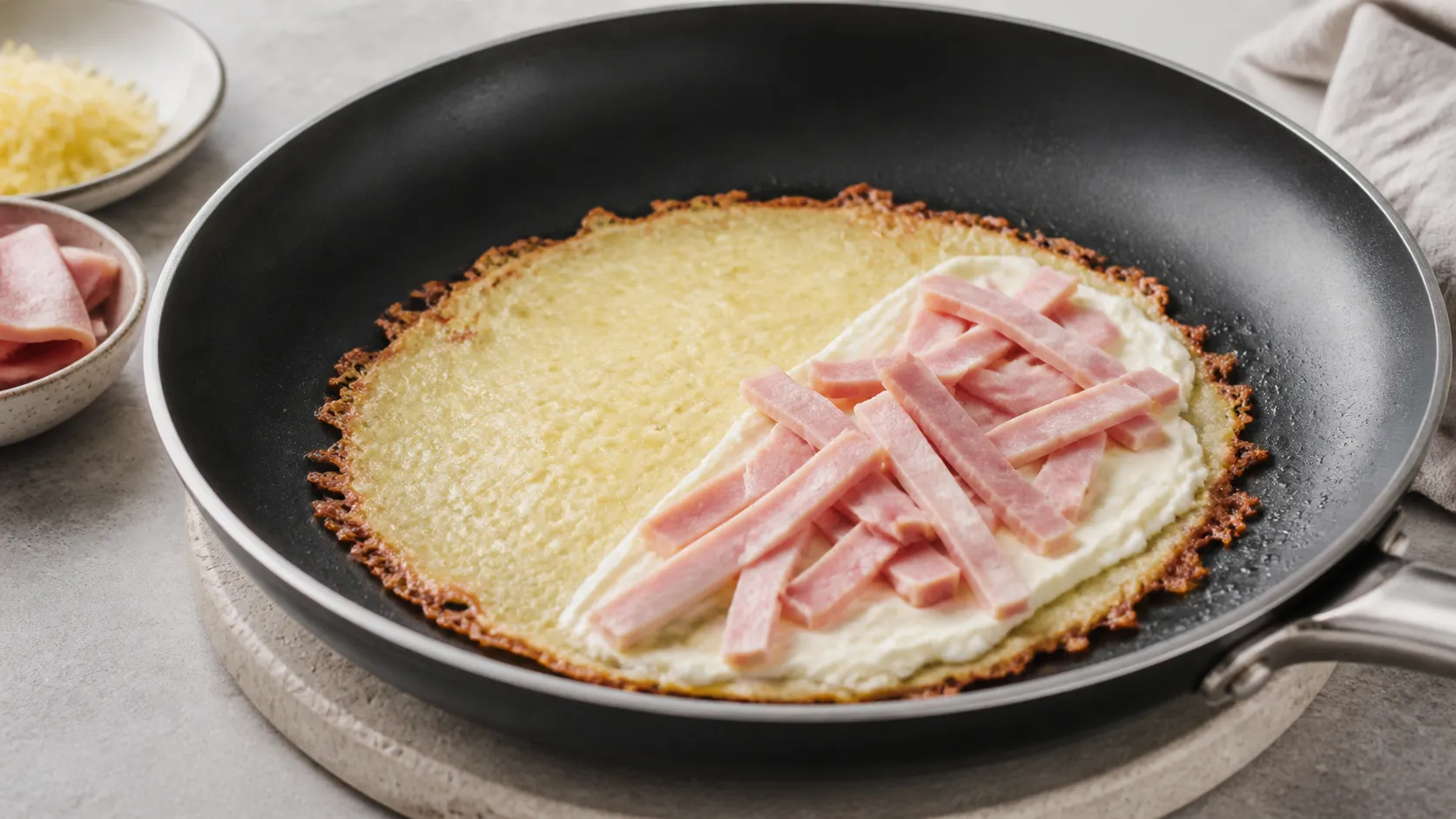 Geschmolzener Käsefladen in der Pfanne, halbseitig mit Frischkäse und Schinken belegt