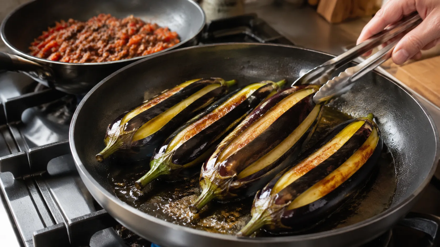 Streifenweise geschälte Auberginen braten goldbraun in einer Pfanne mit Olivenöl, daneben eine Pfanne mit angebratener Rinderhack-Tomaten-Füllung