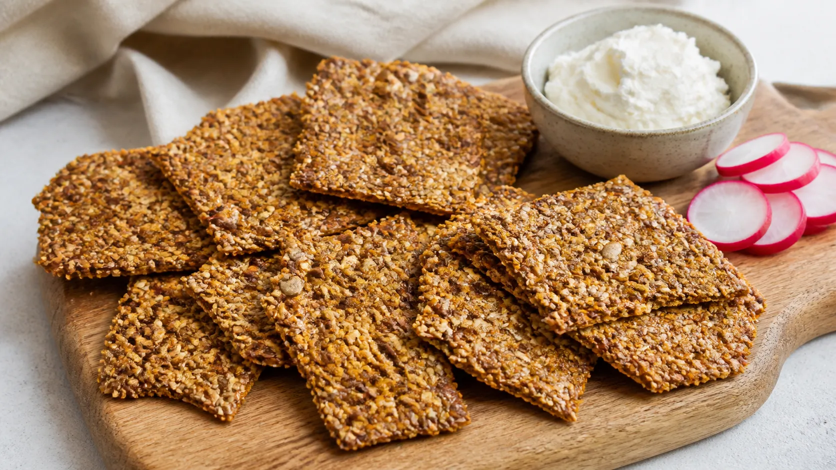 Gebrochene Stücke Keto-Knäckebrot aus Sesam und Leinsamen auf einem Holzbrett, daneben Frischkäse und Radieschen