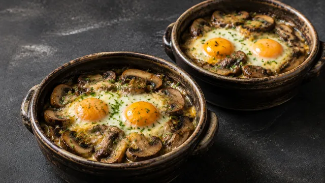 Zwei Auflaufförmchen mit gebackenen Eiern, gebratenen Maronenröhrlingen, Sahne und geschmolzenem Bergkäse, bestreut mit Schnittlauch