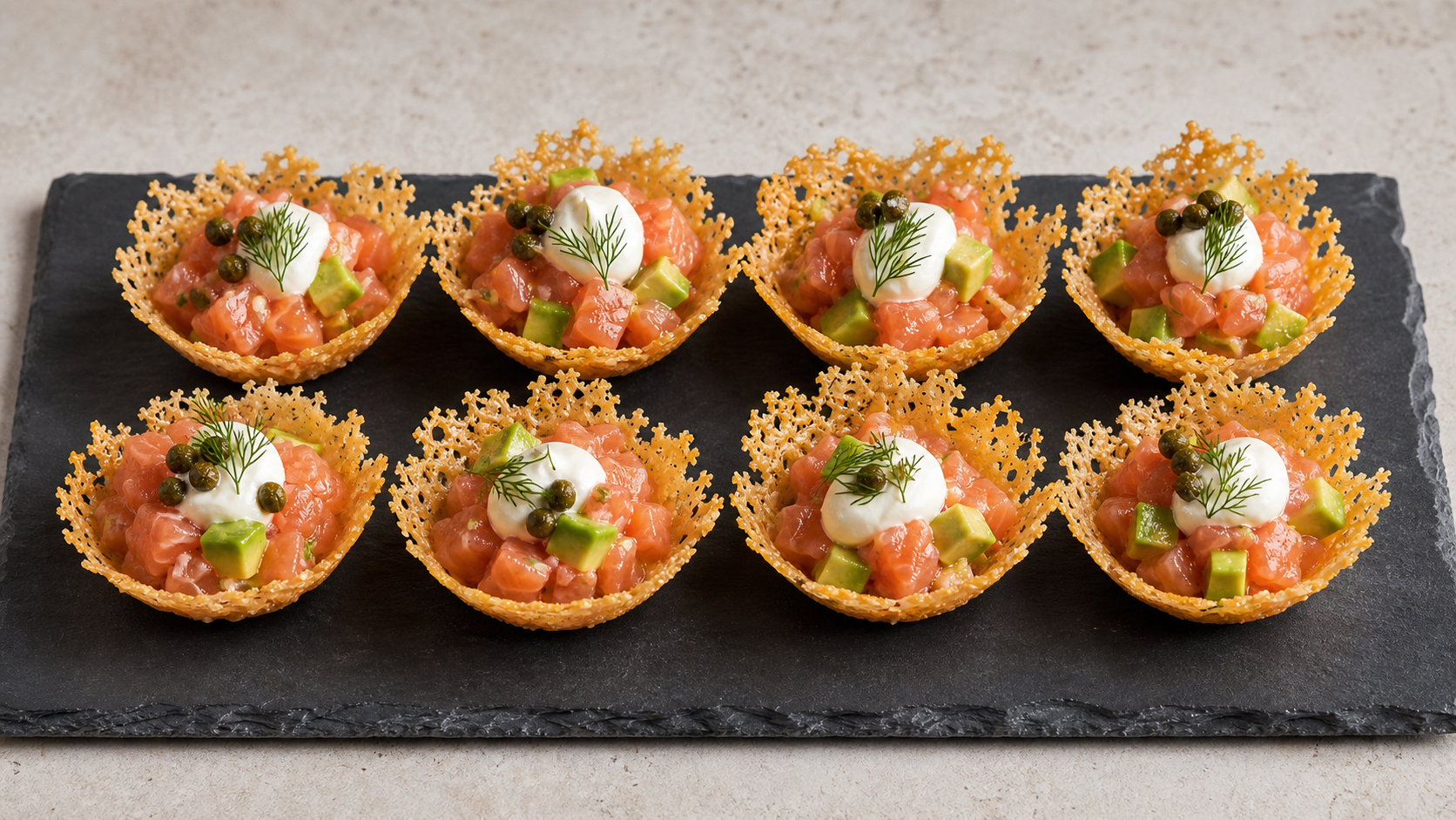 Acht knusprige Parmesan-Körbchen gefüllt mit rosa Lachstatar, Avocadowürfeln, Dillspitzen und Kapern auf einer Schieferplatte