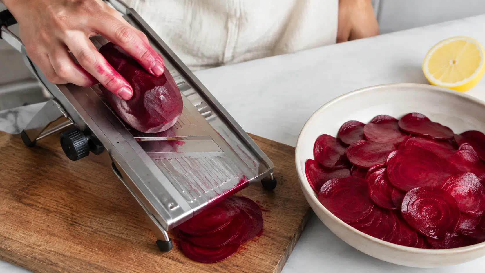 Rote Bete wird auf einem Gemüsehobel in hauchdünne Scheiben geschnitten, daneben eine Schüssel mit den bereits gehobelten Scheiben in Zitronensaft