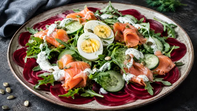 Teller mit hauchdünn gehobelter roher Roter Bete, Rucola, Gurkenscheiben, Räucherlachs, halbiertem hartgekochtem Ei, cremigem weißem Meerrettich-Dressing und gerösteten Kürbiskernen