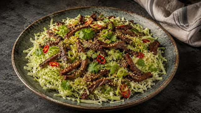 Tiefer Teller mit fein geschnittenem Spitzkohl-Salat, darauf braun angebratene Rindfleischstreifen, gerösteter Sesam, Frühlingszwiebeln und Koriander